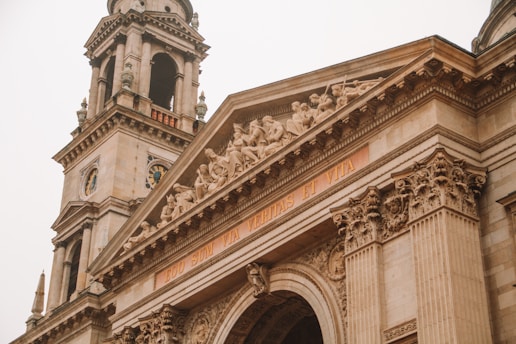 A detailed view of an ornate architectural structure featuring classical elements such as columns and intricate carvings. The facade is adorned with a sculpture depicting several figures in a classical style. Latin text is inscribed below the relief.