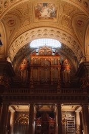 An ornately decorated cathedral interior featuring a grand pipe organ with intricate designs. The ceiling showcases detailed frescoes and gold accents, illuminated by natural light through an arched window.