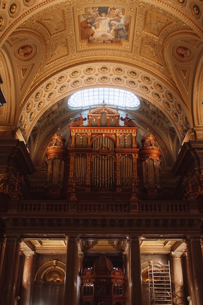 An ornately decorated cathedral interior featuring a grand pipe organ with intricate designs. The ceiling showcases detailed frescoes and gold accents, illuminated by natural light through an arched window.
