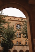 An architectural view captured through an archway, showing a building with intricate brickwork and decorative windows. The building facade features circular and arched window designs with patterned details. Lush green trees partially obscure the building, adding a natural element to the urban scene.