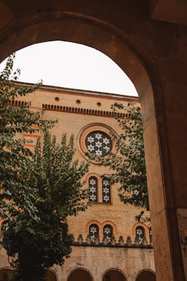 An architectural view captured through an archway, showing a building with intricate brickwork and decorative windows. The building facade features circular and arched window designs with patterned details. Lush green trees partially obscure the building, adding a natural element to the urban scene.