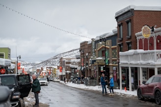 A snowy street scene in a small town with people walking along the sidewalks. The street is lined with brick buildings featuring signs for local businesses like Red Banjo Pizza and the Egyptian Theatre, which has a marquee promoting the Sundance Film Festival. Vehicles are parked along the snowy road, and there are snow-covered rooftops visible against a backdrop of snow-dusted hills.