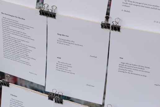 Several sheets of paper clipped together display various poems. The visible pieces contain poems discussing themes such as war, peace, and wishes for a better future. The text is printed in a clean, minimalistic font against a white background, with black clips at the top securing the sheets.