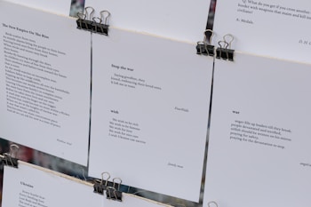 Several sheets of paper clipped together display various poems. The visible pieces contain poems discussing themes such as war, peace, and wishes for a better future. The text is printed in a clean, minimalistic font against a white background, with black clips at the top securing the sheets.
