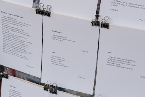 Several sheets of paper clipped together display various poems. The visible pieces contain poems discussing themes such as war, peace, and wishes for a better future. The text is printed in a clean, minimalistic font against a white background, with black clips at the top securing the sheets.