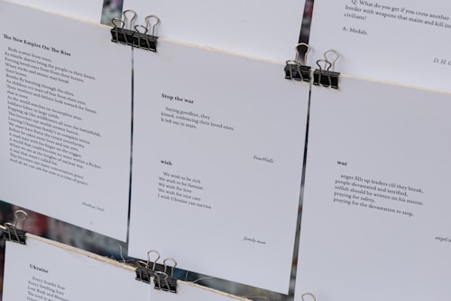 Several sheets of paper clipped together display various poems. The visible pieces contain poems discussing themes such as war, peace, and wishes for a better future. The text is printed in a clean, minimalistic font against a white background, with black clips at the top securing the sheets.