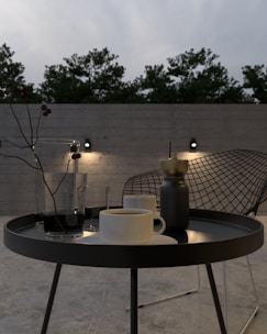 A classic outdoor side table made of weather-resistant metal, holding a lantern and a cup of coffee.