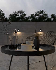 A minimalist outdoor setting featuring a round black metal table with a mug, a glass, and a sleek coffee grinder. In the background, there is a modern wire chair and a concrete wall with mounted lights, all surrounded by trees under an overcast sky.