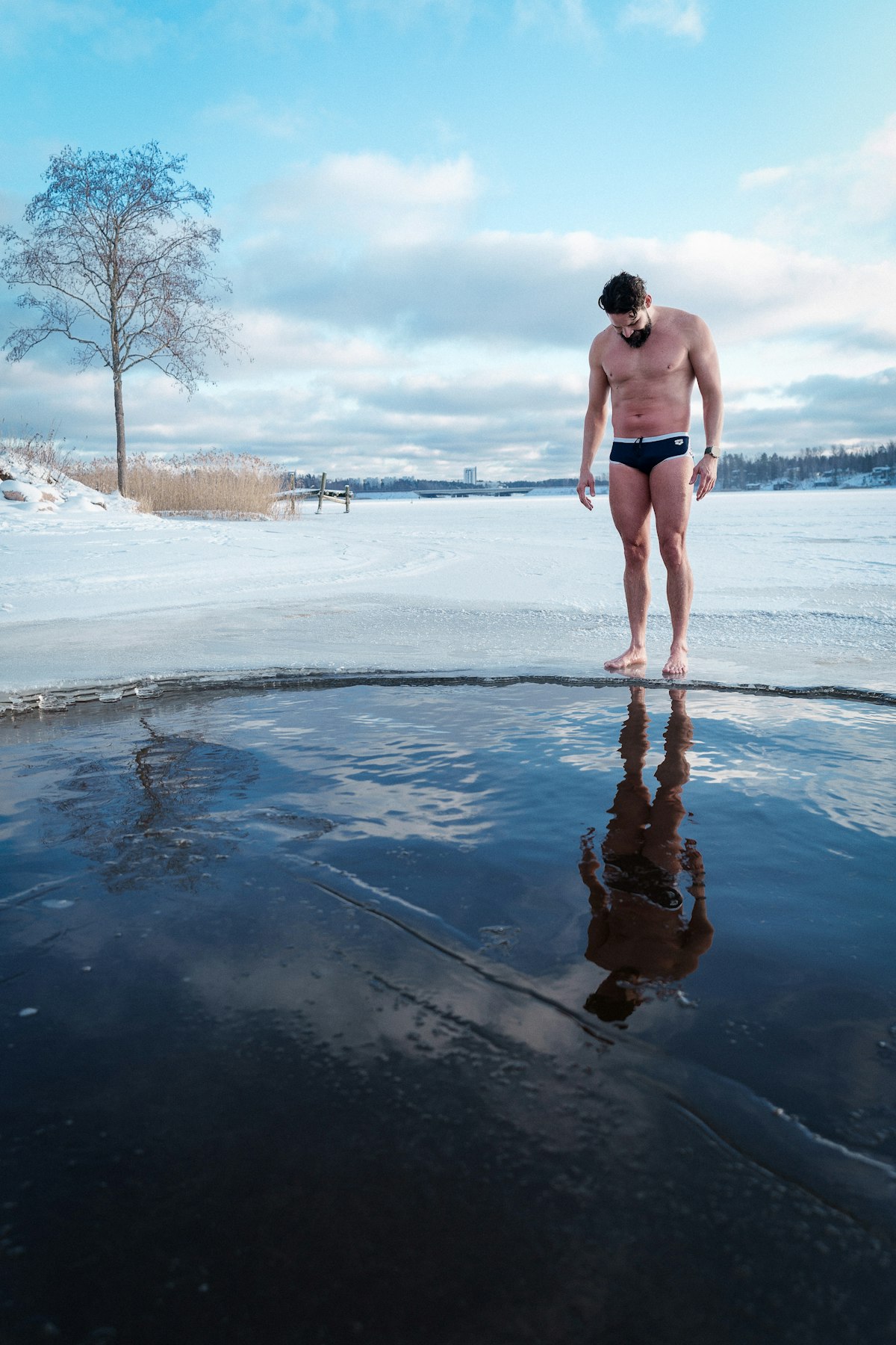 Solo figure standing barefoot beside an ice hole on a frozen Finnish lake under winter light