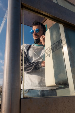 A man wearing blue sunglasses and a white shirt is making a call from a vintage phone booth. The booth is metallic with glass panels, and the telephone has a coiled cord. The scene is captured in daylight, with a clear blue sky visible in the background.
