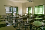 An empty classroom with rows of desks and chairs arranged neatly. Green curtains partially cover the windows, allowing some natural light to seep in. The floor is tiled in a checkered pattern of light and dark green, matching the color scheme of the furniture. A ceiling-mounted projector is visible above the desks.