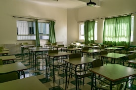 An empty classroom with rows of desks and chairs arranged neatly. Green curtains partially cover the windows, allowing some natural light to seep in. The floor is tiled in a checkered pattern of light and dark green, matching the color scheme of the furniture. A ceiling-mounted projector is visible above the desks.