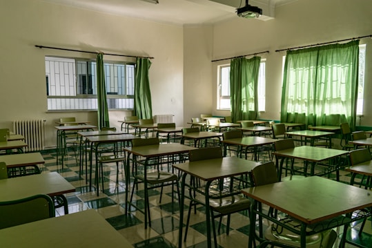 An empty classroom with rows of desks and chairs arranged neatly. Green curtains partially cover the windows, allowing some natural light to seep in. The floor is tiled in a checkered pattern of light and dark green, matching the color scheme of the furniture. A ceiling-mounted projector is visible above the desks.