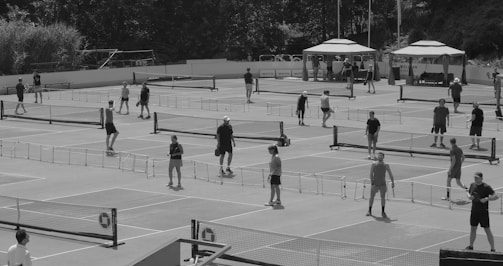 Multiple people are actively engaged in playing pickleball on several outdoor courts. The setting includes surrounding foliage and tents likely for spectators or check-in areas.