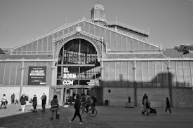 The image depicts a large historical structure with elaborate ironwork and glass design, featuring a prominent entrance marked with the words 'El Born CCM'. Several people are walking, standing, and riding bicycles in front of the building, indicating a lively urban setting.