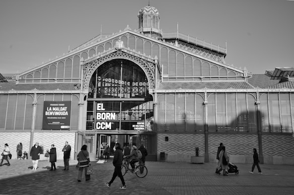 The image depicts a large historical structure with elaborate ironwork and glass design, featuring a prominent entrance marked with the words 'El Born CCM'. Several people are walking, standing, and riding bicycles in front of the building, indicating a lively urban setting.