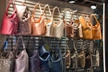 A display of handbags arranged in two rows inside a store window. The upper row features bags in warm tones of pink, peach, beige, and tan, while the lower row includes bags in cooler shades such as purple, black, and brown. The bags are well-lit, highlighting their textures and materials, with a light cream background of horizontal slats.