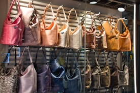 A display of handbags arranged in two rows inside a store window. The upper row features bags in warm tones of pink, peach, beige, and tan, while the lower row includes bags in cooler shades such as purple, black, and brown. The bags are well-lit, highlighting their textures and materials, with a light cream background of horizontal slats.
