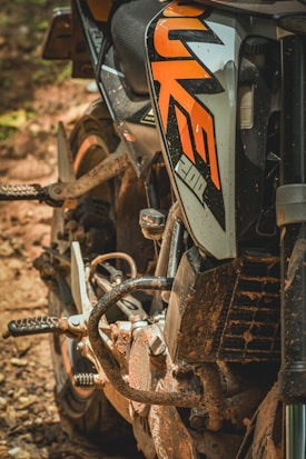 A close-up view of a motorcycle side with a well-defined orange logo or decal. The bike appears somewhat dirty, indicating off-road or rugged usage. The tires and metal parts are also visible, covered in mud and dust.