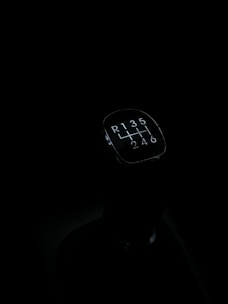 A gear shift knob of a manual transmission vehicle is seen against a dark background. The knob displays the shift pattern, which includes reverse and gears 1 through 6.