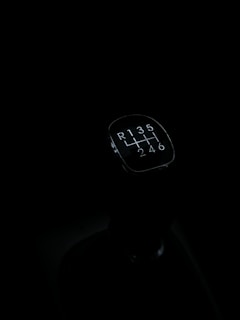 A gear shift knob of a manual transmission vehicle is seen against a dark background. The knob displays the shift pattern, which includes reverse and gears 1 through 6.
