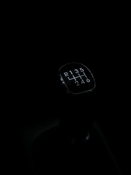 A gear shift knob of a manual transmission vehicle is seen against a dark background. The knob displays the shift pattern, which includes reverse and gears 1 through 6.