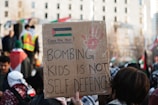 A person is holding a cardboard sign at a protest. The sign, written in bold letters, reads 'Bombing kids is not self defence.' The sign also contains a red handprint and a drawing resembling the Palestinian flag with the words 'Cease Fire Now!' written above it. The background shows other protestors and flags, with a cityscape scene comprising a tall building with multiple windows.