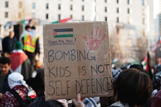 A person is holding a cardboard sign at a protest. The sign, written in bold letters, reads 'Bombing kids is not self defence.' The sign also contains a red handprint and a drawing resembling the Palestinian flag with the words 'Cease Fire Now!' written above it. The background shows other protestors and flags, with a cityscape scene comprising a tall building with multiple windows.