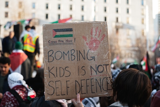 A person is holding a cardboard sign at a protest. The sign, written in bold letters, reads 'Bombing kids is not self defence.' The sign also contains a red handprint and a drawing resembling the Palestinian flag with the words 'Cease Fire Now!' written above it. The background shows other protestors and flags, with a cityscape scene comprising a tall building with multiple windows.