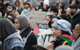 A diverse crowd is gathered, holding signs with messages of unity and support for Palestine. Many individuals wear winter clothing, with some wearing face masks. The signs are prominently displayed, indicating themes of solidarity.