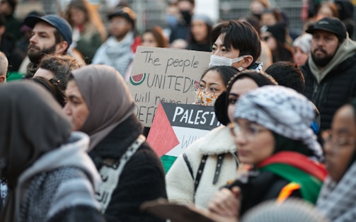 A diverse crowd is gathered, holding signs with messages of unity and support for Palestine. Many individuals wear winter clothing, with some wearing face masks. The signs are prominently displayed, indicating themes of solidarity.