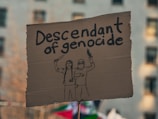A cardboard sign with the words 'Descendant of genocide' written in black marker. Below the text, there are hand-drawn illustrations of two individuals, one holding a knife and the other holding what appears to be a plant or branch. The background is blurry, indicating an outdoor setting, possibly during a protest or demonstration.