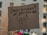 A cardboard sign with the words 'Descendant of genocide' written in black marker. Below the text, there are hand-drawn illustrations of two individuals, one holding a knife and the other holding what appears to be a plant or branch. The background is blurry, indicating an outdoor setting, possibly during a protest or demonstration.
