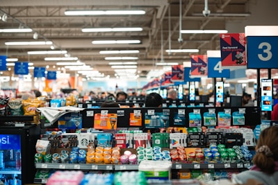 A busy supermarket checkout area with various products displayed at the counter, including snacks, beverages, magazines, and gift cards. The scene shows multiple checkout lanes with signs numbered three and four visible, and a group of shoppers waiting in line. The store's bright, organized atmosphere features overhead lighting and shelves filled with colorful items.