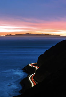 A cinematic shot of a van winding along a coastal highway with cliffs and ocean waves under warm sunset light.
