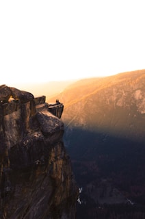 A breathtaking landscape featuring a high cliff with rocky edges. Two people sit at the edge of the cliff, overlooking a vast valley filled with trees. The sky is clear, and the warm glow of the setting sun casts a golden hue over the scene, emphasizing the rugged texture of the rock and the distant mountains.