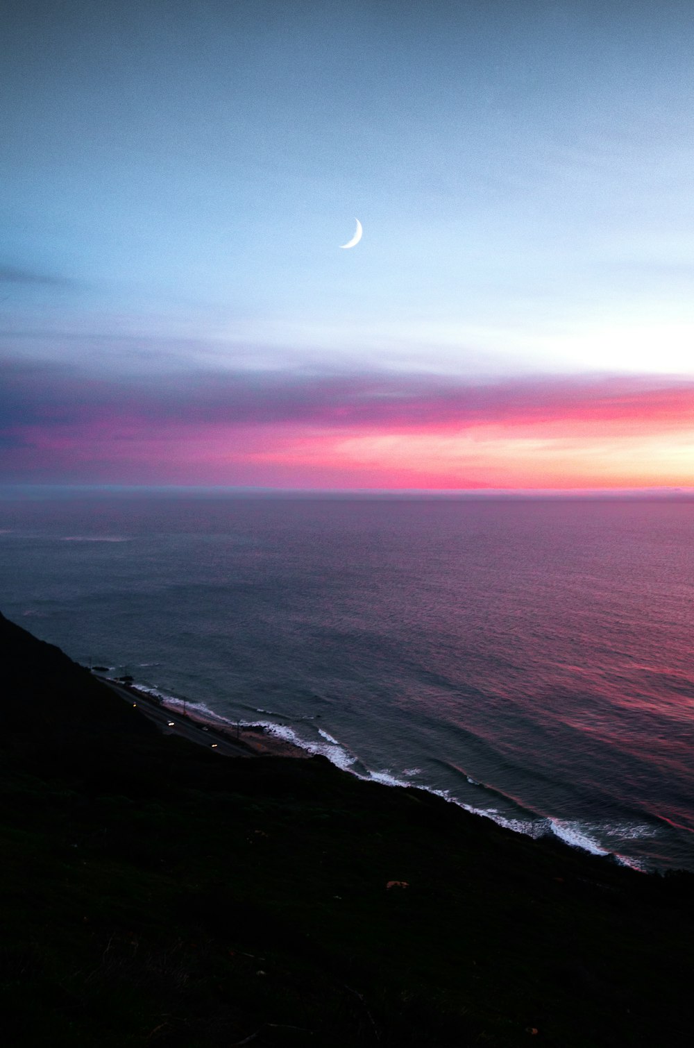 a sunset over the ocean with a half moon in the sky