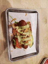 A loaded hot dog is placed on a rectangular metal tray. The hot dog bun is topped with sausages, slices of tomato, avocado, crispy fried onions, and a drizzle of green sauce. The tray is lined with a paper that has printed text on it.