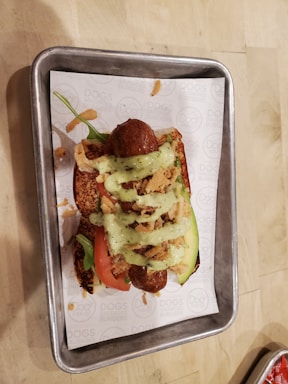 A loaded hot dog is placed on a rectangular metal tray. The hot dog bun is topped with sausages, slices of tomato, avocado, crispy fried onions, and a drizzle of green sauce. The tray is lined with a paper that has printed text on it.