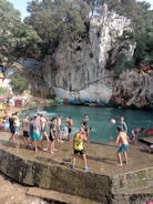 A group of people are gathered around a natural swimming area surrounded by rocky cliffs and trees. Some individuals are playing with a ball, while others are standing or sitting near the water. Colorful chairs are placed around the area, and the atmosphere is lively with people enjoying leisure time.