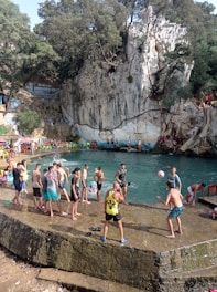 A group of people are gathered around a natural swimming area surrounded by rocky cliffs and trees. Some individuals are playing with a ball, while others are standing or sitting near the water. Colorful chairs are placed around the area, and the atmosphere is lively with people enjoying leisure time.