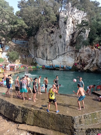 A group of people are gathered around a natural swimming area surrounded by rocky cliffs and trees. Some individuals are playing with a ball, while others are standing or sitting near the water. Colorful chairs are placed around the area, and the atmosphere is lively with people enjoying leisure time.