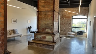 An industrial-style room featuring exposed brick walls, large windows, and modern furniture. The space includes a large brick column in the center, a couch with pillows, and a sleek black chair. Overhead, long LED lights are suspended from the dark ceiling, creating a minimalist and stylish atmosphere.