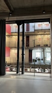 A modern interior space characterized by industrial design elements, including visible steel beams and pillars. The multi-level structure features balconies with plants growing along wire mesh railings. The walls display large red numerals indicating room numbers, and large windows allow for natural lighting. Hanging lights are minimalist, contributing to the industrial aesthetic.