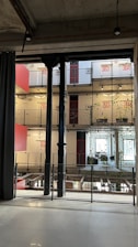 A modern interior space characterized by industrial design elements, including visible steel beams and pillars. The multi-level structure features balconies with plants growing along wire mesh railings. The walls display large red numerals indicating room numbers, and large windows allow for natural lighting. Hanging lights are minimalist, contributing to the industrial aesthetic.
