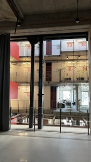 A modern interior space characterized by industrial design elements, including visible steel beams and pillars. The multi-level structure features balconies with plants growing along wire mesh railings. The walls display large red numerals indicating room numbers, and large windows allow for natural lighting. Hanging lights are minimalist, contributing to the industrial aesthetic.
