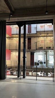 A modern interior space characterized by industrial design elements, including visible steel beams and pillars. The multi-level structure features balconies with plants growing along wire mesh railings. The walls display large red numerals indicating room numbers, and large windows allow for natural lighting. Hanging lights are minimalist, contributing to the industrial aesthetic.