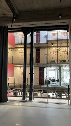 A modern interior space characterized by industrial design elements, including visible steel beams and pillars. The multi-level structure features balconies with plants growing along wire mesh railings. The walls display large red numerals indicating room numbers, and large windows allow for natural lighting. Hanging lights are minimalist, contributing to the industrial aesthetic.
