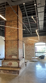 A modern industrial interior with exposed brickwork and a large central brick column. The room features high ceilings with visible metal beams and suspended linear lighting. There are large windows allowing natural light to fill the space, alongside minimalistic furniture such as a small table and chairs by the window.