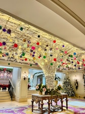 A festive indoor scene with a large, intricately decorated tree-like structure adorned with numerous colorful ornaments and sparkling lights. The room features ornate furniture, elegant chandeliers, and additional Christmas decorations such as garlands and a Christmas tree in the background.